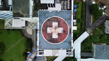 This footage presents an aerial, top-down view of a hospital helipad marked with a red cross. The helipad is surrounded by buildings, green lawns, trees, and a parking area. - Powered by Adobe
