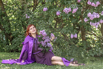 A beautiful young woman with red hair, against a background of blooming lilacs, in a cloak and hood. Medieval fantasy costume. Fairy tale heroine, princess, duchess.