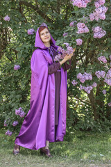 A beautiful young woman with red hair, against a background of blooming lilacs, in a cloak and hood. Medieval fantasy costume. Fairy tale heroine, princess, duchess.