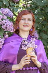 A beautiful young woman with red hair, against a background of blooming lilacs, in a cloak and hood. Medieval fantasy costume. Fairy tale heroine, princess, duchess.