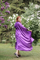 A beautiful young woman with red hair, against a background of blooming lilacs, in a cloak and hood. Medieval fantasy costume. Fairy tale heroine, princess, duchess.