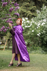 A beautiful young woman with red hair, against a background of blooming lilacs, in a cloak and hood. Medieval fantasy costume. Fairy tale heroine, princess, duchess.