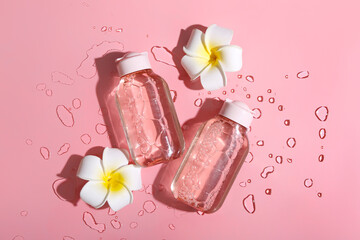 Bottles of micellar water, plumeria flowers and water drops on pink background, closeup