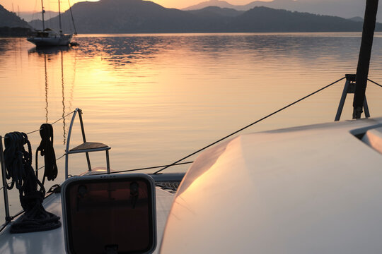 scenic view of morning calm tranquil sunrise from the boat deck at anchor stop, rising sun, mountains, sea with light reflections, twilight colors, sailing boats, serene harbour