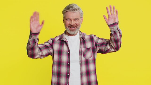 Happy Caucasian man smiling friendly at camera, waving hands gesturing hello greeting or goodbye welcoming with invitation hospitable expression. Handsome guy isolated on yellow studio background