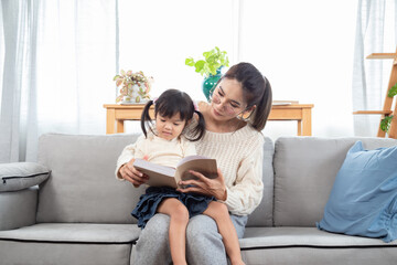Fototapeta premium Asian beautiful pregnant mother reads a book with her eldest daughter in the living room during their leisure time, mom teaching baby to read, Telling stories to little child. family happiness concept