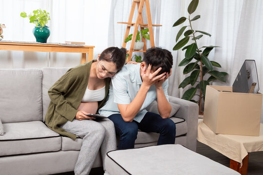 Pregnant woman sitting on couch, comforting stressed boyfriend holding head in hands, depicts emotional support, job loss stress, and challenges faced by expecting parents.