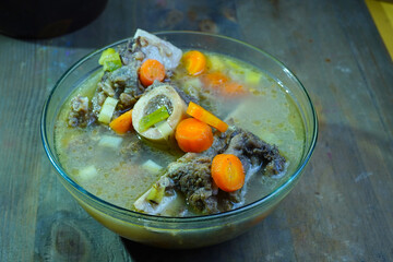 A close-up of a hearty bone marrow soup, featuring tender meat, vibrant carrots, and green vegetables in a clear glass bowl on a rustic wooden table.