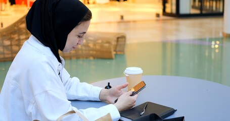 Young muslim woman wearing traditional hijab sitting at a table in a shopping mall, using her smartphone with shopping bags and a coffee next to her. Happy Muslim woman in black hijab
