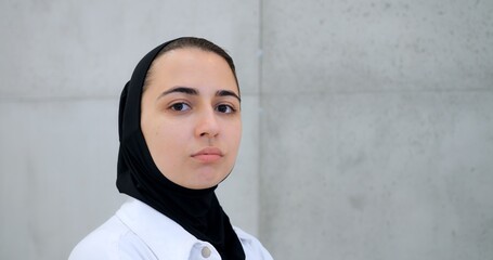 Professional muslim female healthcare worker wearing hijab and white coat, standing against concrete wall, displaying confident facial expressions while facing camera