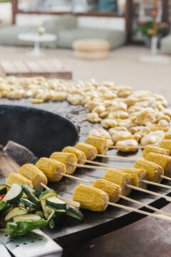 Assorted vegetables and corn on skewers grilling on a round fire plate during an outdoor cooking event.