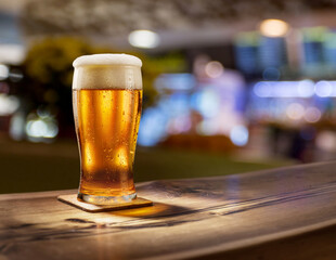 Glass of chilled beer on wooden bar tabletop and blurred brightly lit bar interior in the background.