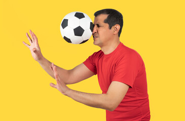 Man with red t-shirt playing sport being hit by a soccer ball with force in the face when he misses a catch or as an unexpected accident to a spectator,against a yellow background © cunaplus
