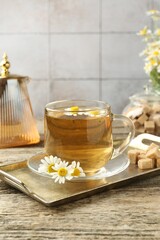 Delicious chamomile tea in glass cup, fresh flowers, brown sugar and teapot on wooden table, closeup