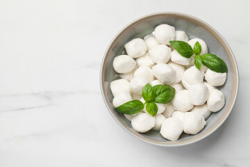 Tasty mozzarella cheese balls in bowl and basil on white marble table, top view. Space for text