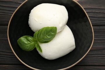 Pieces of delicious mozzarella cheese and basil in bowl on wooden table, top view