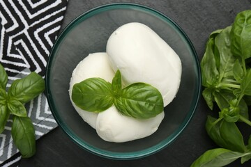 Pieces of delicious mozzarella cheese and basil in bowl on dark textured table, flat lay