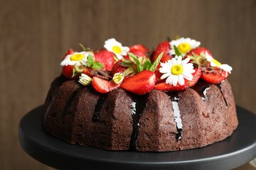 Delicious chocolate bundt cake with strawberries, chamomile flowers and mint on stand against blurred brown background, closeup