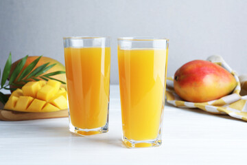 Tasty mango juice in glasses, fresh fruits and green leaves on white wooden table