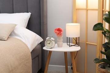 Side table with lamp, alarm clock and flower near bed indoors