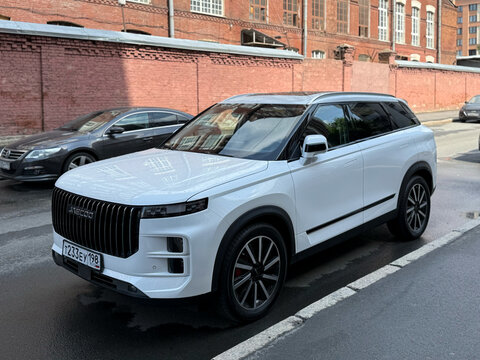 A modern white Jaecoo J7 luxury crossover car parked on a city street pavement against a red brick wall background.