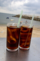 glass of iced cola sits on a table by the sea, with a cloudy sky backdrop. The drink's bubbles and condensation are visible