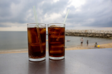 glass of iced cola sits on a table by the sea, with a cloudy sky backdrop. The drink's bubbles and condensation are visible