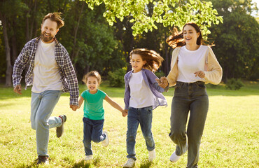 Fototapeta premium Happy family running and having fun in summer park enjoying sunny day in nature. Mother, father and two cheerful children boy and girl walking on green grass outdoors at sunset together.