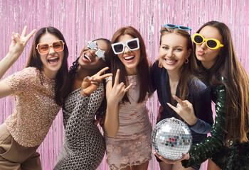Group of five smiling diverse stylish young women posing at party. Cheerful female friends wearing funky sunglasses and festive outfits making group photo and having fun against shiny pink backdrop.