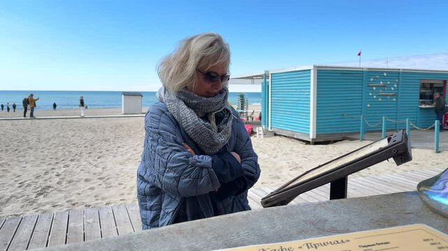 A woman in a jacket looks at the menu outside a beachside restaurant on a cold day. The scene captures a quiet seaside atmosphere with cool weather and a casual, contemplative moment