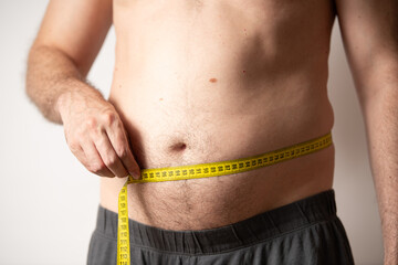 A man stands in a casual setting, measuring his waist with a tape measure. He is focused on tracking his fitness progress while dressed in comfortable shorts. It is evening time