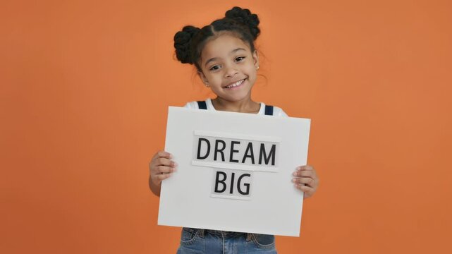 Happy girl holding dream big text sign on orange background, inspiring ambition, confidence, and hope in the next generation