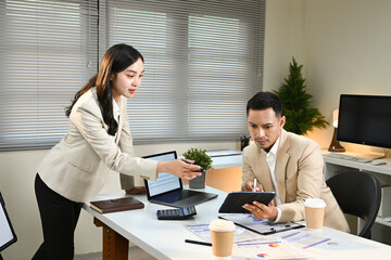 A woman and a man are sitting at a desk with a laptop and a tablet. The woman is handing the tablet to the man, possibly showing him something on the screen