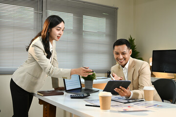 A woman and a man are sitting at a desk with a laptop and a tablet. The woman is handing the tablet to the man, possibly showing him something on the screen