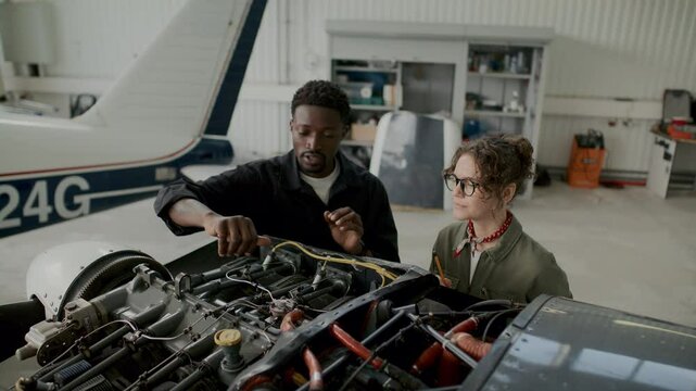 African American mechanic and his Caucasian female colleague discussing components of open aircraft engine while working together inside aviation hangar - Powered by Adobe