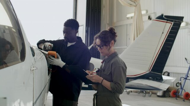 African American avionics technician using digital multimeter and talking to Caucasian female colleague taking notes during aircraft repair work in maintenance hangar