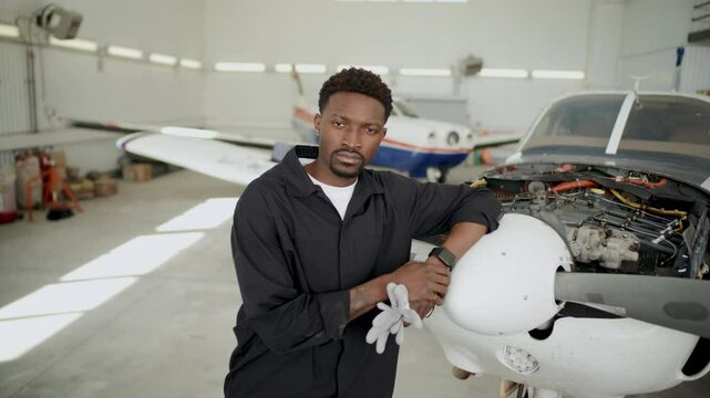 Young Black male aircraft mechanic in workwear standing beside plane with open engine compartment in hangar, holding gloves and looking confidently into camera. Video portrait
