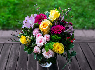 Bunter Blumenstrauß mit gelben Rosen, rosa Rosen und Gerbera auf einem Holztisch mit  grüner Wiese im Hintergrund