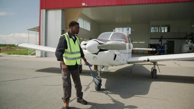 African American technician in reflective vest manually towing light aircraft from hangar using tow bar on sunny day at airfield