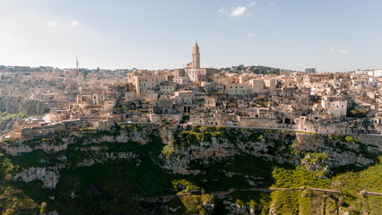 Fototapeta premium Aerial view of Matera, Italy, with its stone-built houses and cathedral tower rising above the deep ravine. A bright, clear day highlights the timeless beauty of this ancient city