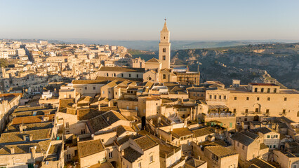 Aerial view of Matera, Italy, bathed in golden morning light. The iconic stone dwellings and cathedral tower rise above the canyon, showcasing this UNESCO-listed historic wonder