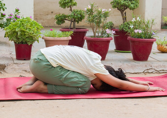 Asian little girl wearing track pant and t-shirts doing balasana yoga pose on mat outside the house. healthy meditation for kids mental health.