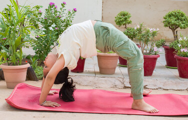 A flexible young girl performs Urdhva Dhanurasana, also known as the upward bow or wheel yoga pose. wearing track pants and a t-shirt in a lush green garden.