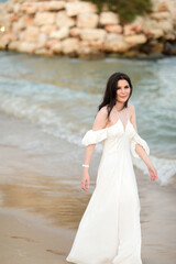 Beautiful young girl with long dark hair in a white long dress on the beach at sea in summer