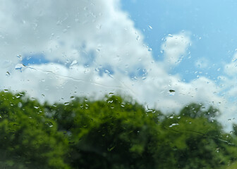 Raindrops on a car window reflect a sunny sky with clouds above lush green trees