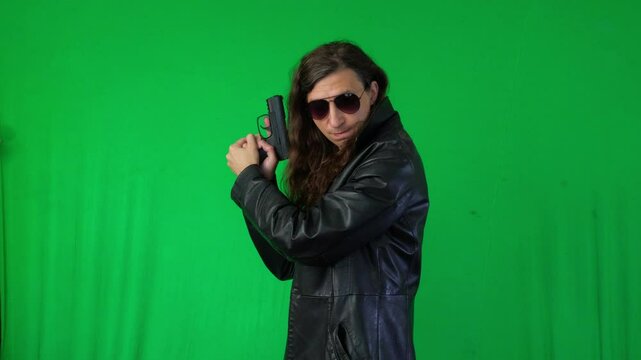 A mid-shot captures a serious and ready mood as a man in a leather jacket and sunglasses holds a handgun with both hands, looking directly at the camera, against a green screen