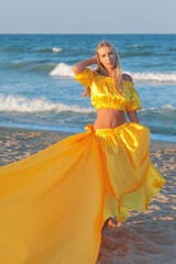 Beautiful young girl with long blonde hair in a yellow long dress with a train on the beach at sea...