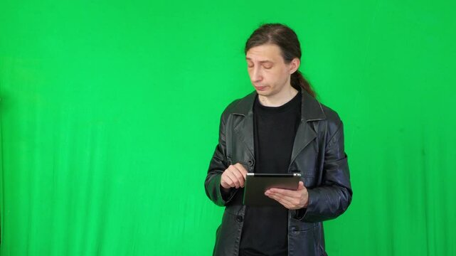 A mid-shot captures a focused and modern mood as a man in a black leather jacket intently uses a digital tablet, suggesting technology interaction or data review, against a green screen background