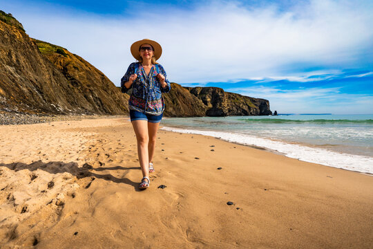 Middle aged woman in sun hat walking towards cliffs on sandy beach and enjoying holidays on spring day. Arrifana Praia, Portugal. Front view