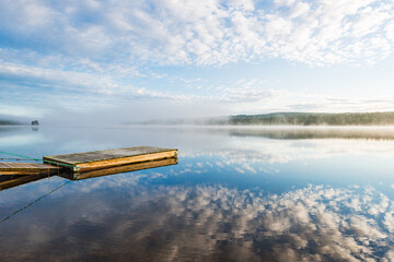 Serene jetty on a still lake during early morning mist in Sweden showcases reflections of the sky
